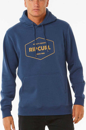 A person is modeling a blue Rip Curl Stapler Hood, featuring a yellow logo that reads "Quality Goods Rip Curl Since 1969" on the front. This Cotton Polyester Fleece Hoodie comes with a front pocket and drawstrings, set against a plain white background.