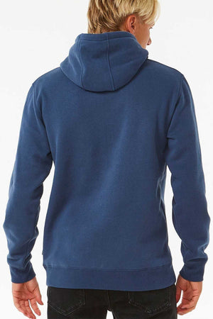 A blond-haired person facing away from the camera wears a blue Rip Curl Stapler Hood and black pants. The image highlights the hoodie’s back, hood, and long sleeves against a white backdrop.