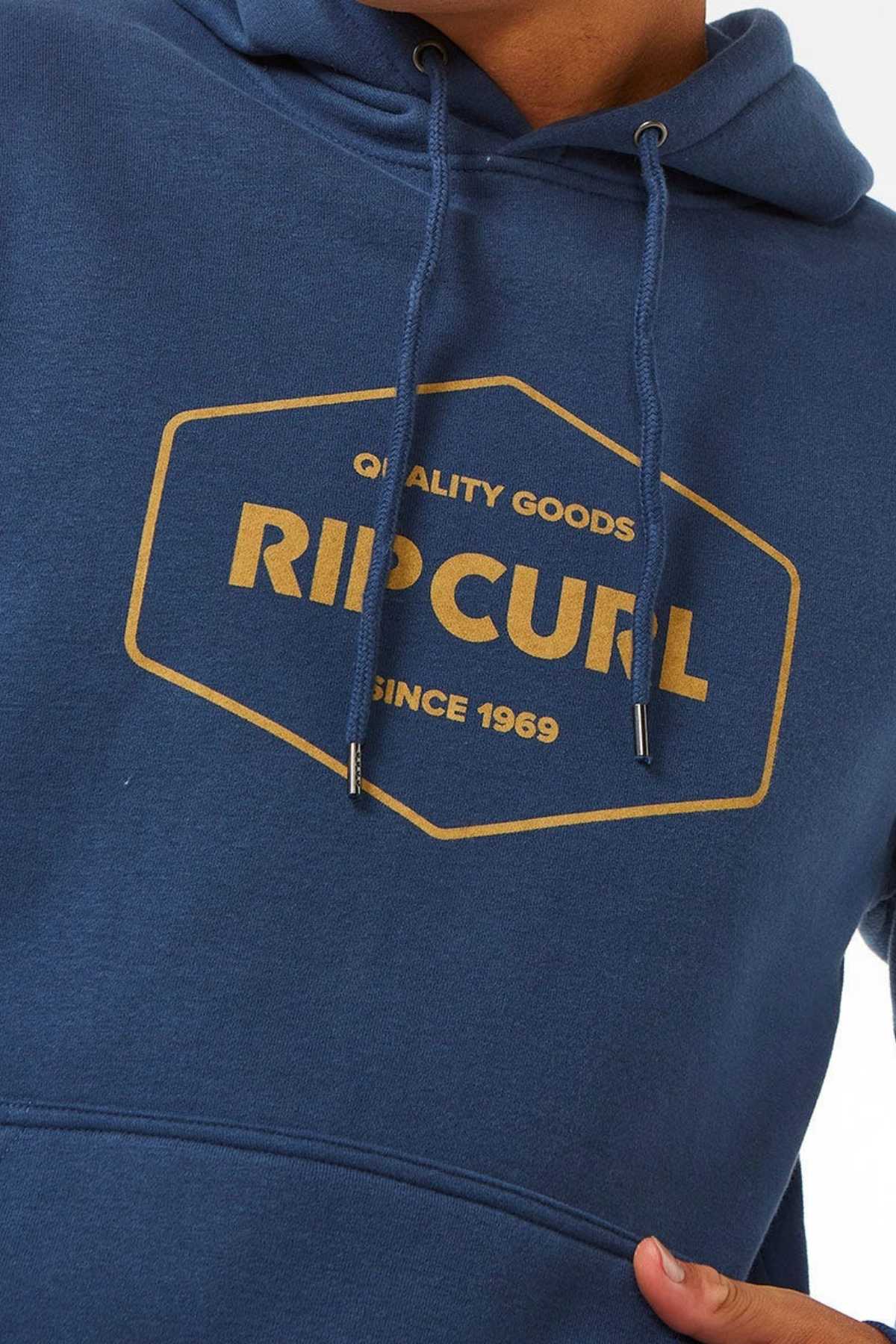 A person wearing a Rip Curl Stapler Hood, a blue hoodie made of cotton polyester fleece, showcases "Quality Goods Rip Curl Since 1969" in a yellow hexagon-shaped print on the front.