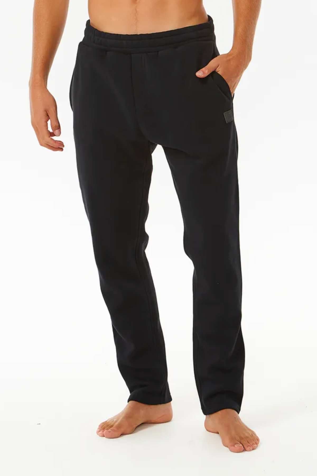 A person stands barefoot wearing Rip Curl Stapler Trackpant joggers with an elastic waist and hands in pockets, against a white background. The top half of the body is not visible.