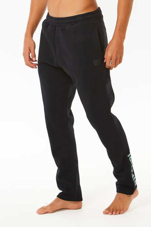 A person stands barefoot wearing Rip Curl Stapler Trackpant in black, featuring an elastic waist, Rip Curl logo on the lower left leg, side pockets, and a soft fleece lining. The background is plain white.