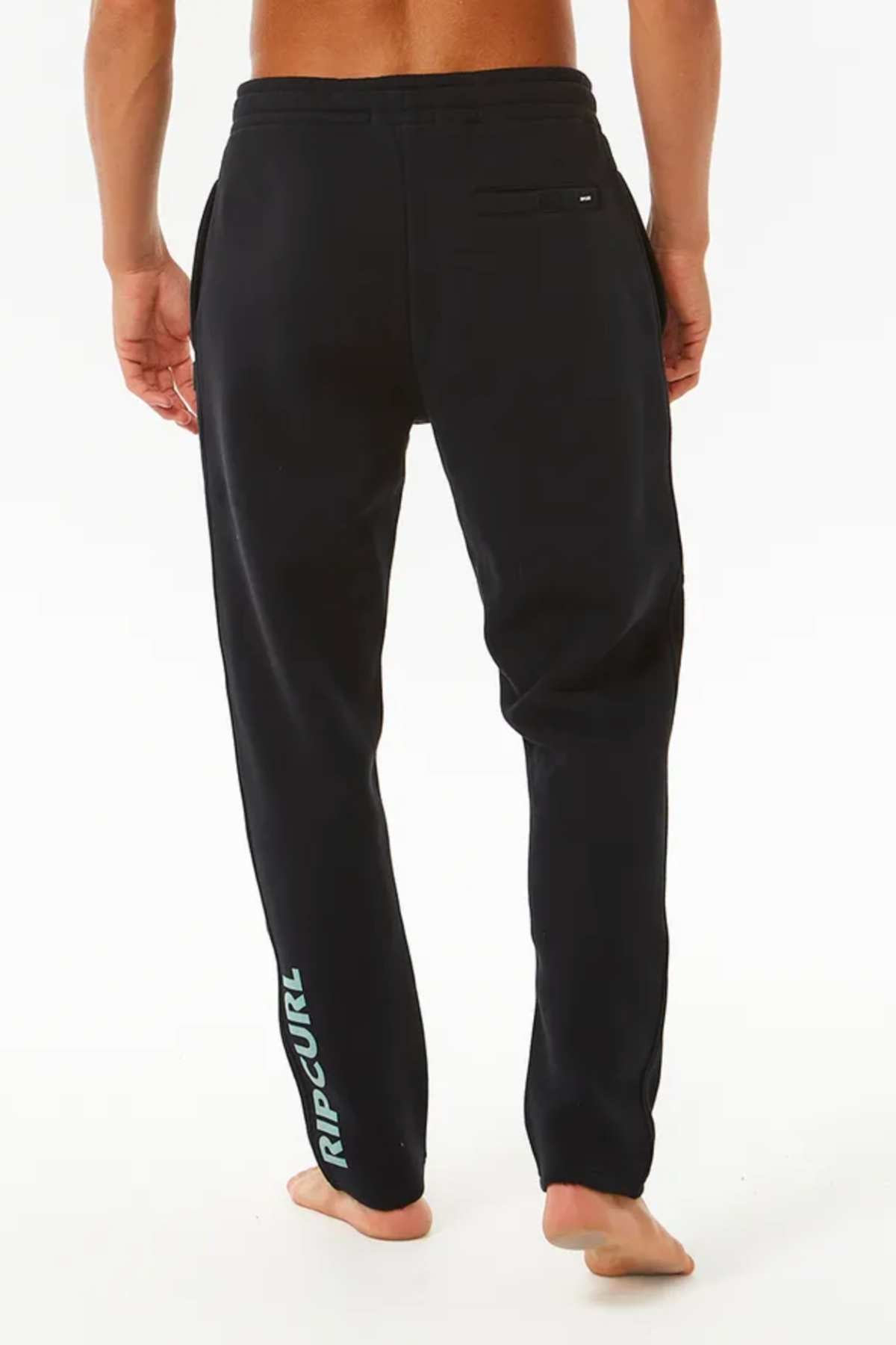 A shirtless person stands barefoot facing away, wearing Rip Curl Stapler Trackpant in black with an elastic waist, side and back pockets, and "Rip Curl" in white and teal on the lower left leg. Features the iconic Rip Curl logo.