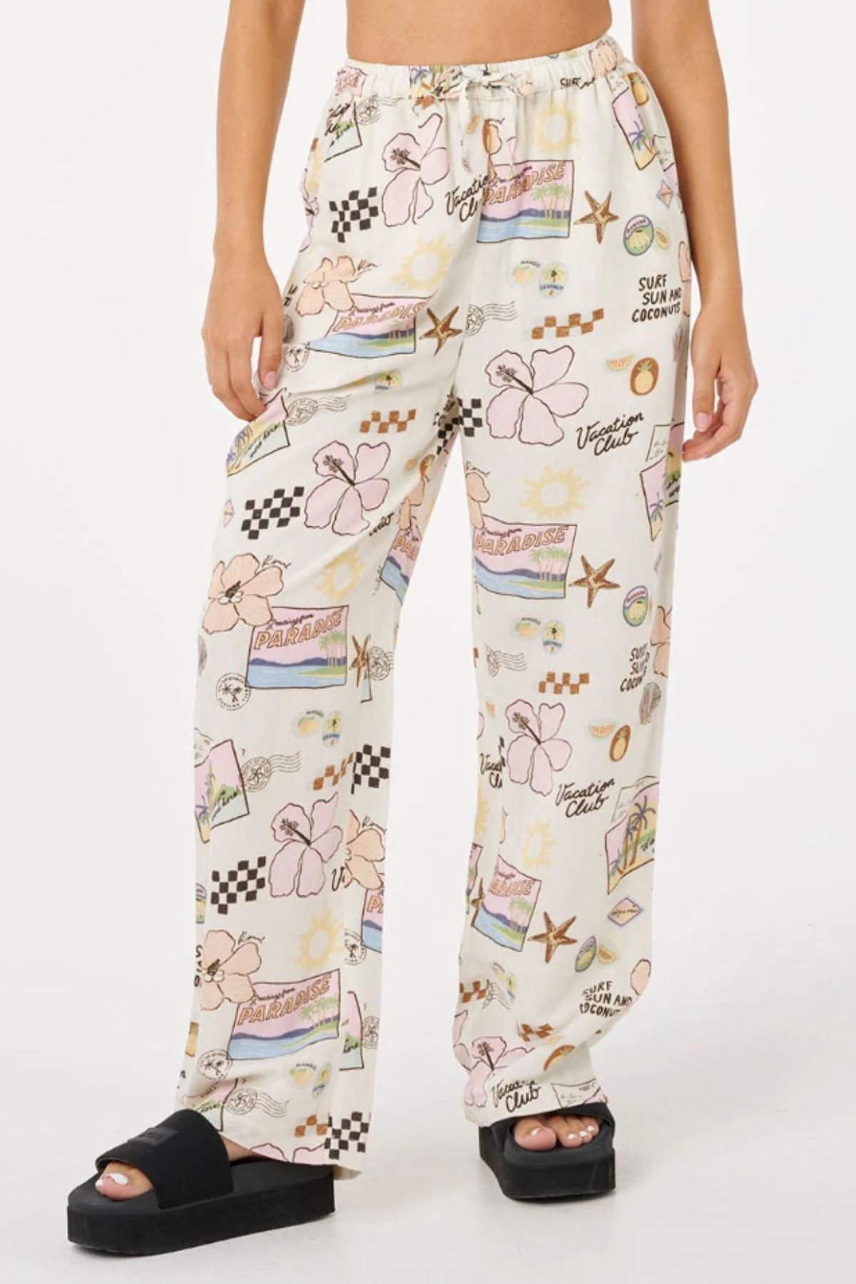 A person wears Rip Curl Sunset Luau Wide Leg Pants in a linen-viscose blend with vibrant summer prints, styled with black platform sandals. The warm-weather look pops against the plain white background.