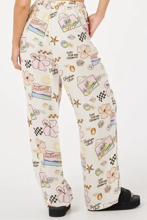 A person wears the Rip Curl Sunset Luau Wide Leg Pant, featuring a loose white fit with colorful tropical prints of palm trees, flowers, starfish, shells, and beach-themed words—ideal for warm-weather style.