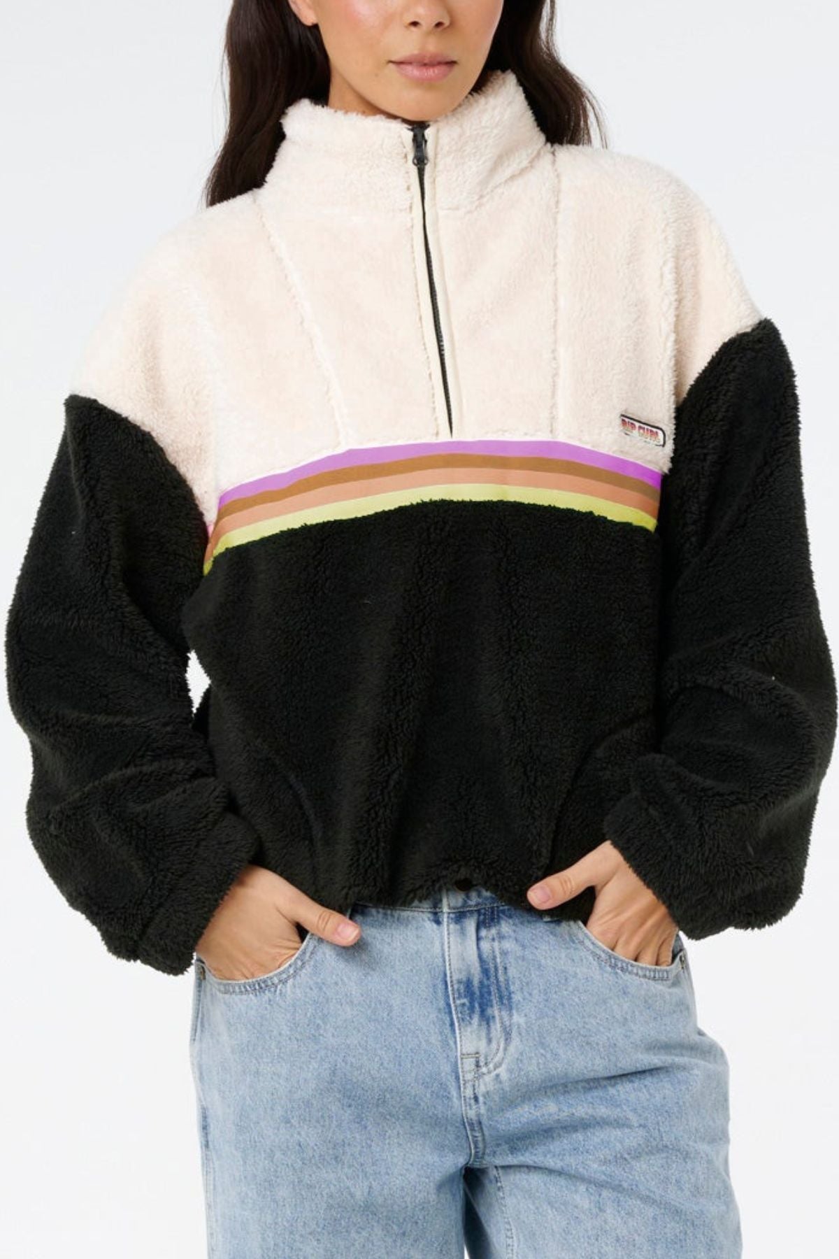 A person is wearing the Rip Curl Surf Revival Polar Fleece, a fuzzy half-zip pullover with a white top, black bottom, and horizontal pink, yellow, and green stripes across the chest; their hands are in their pockets.