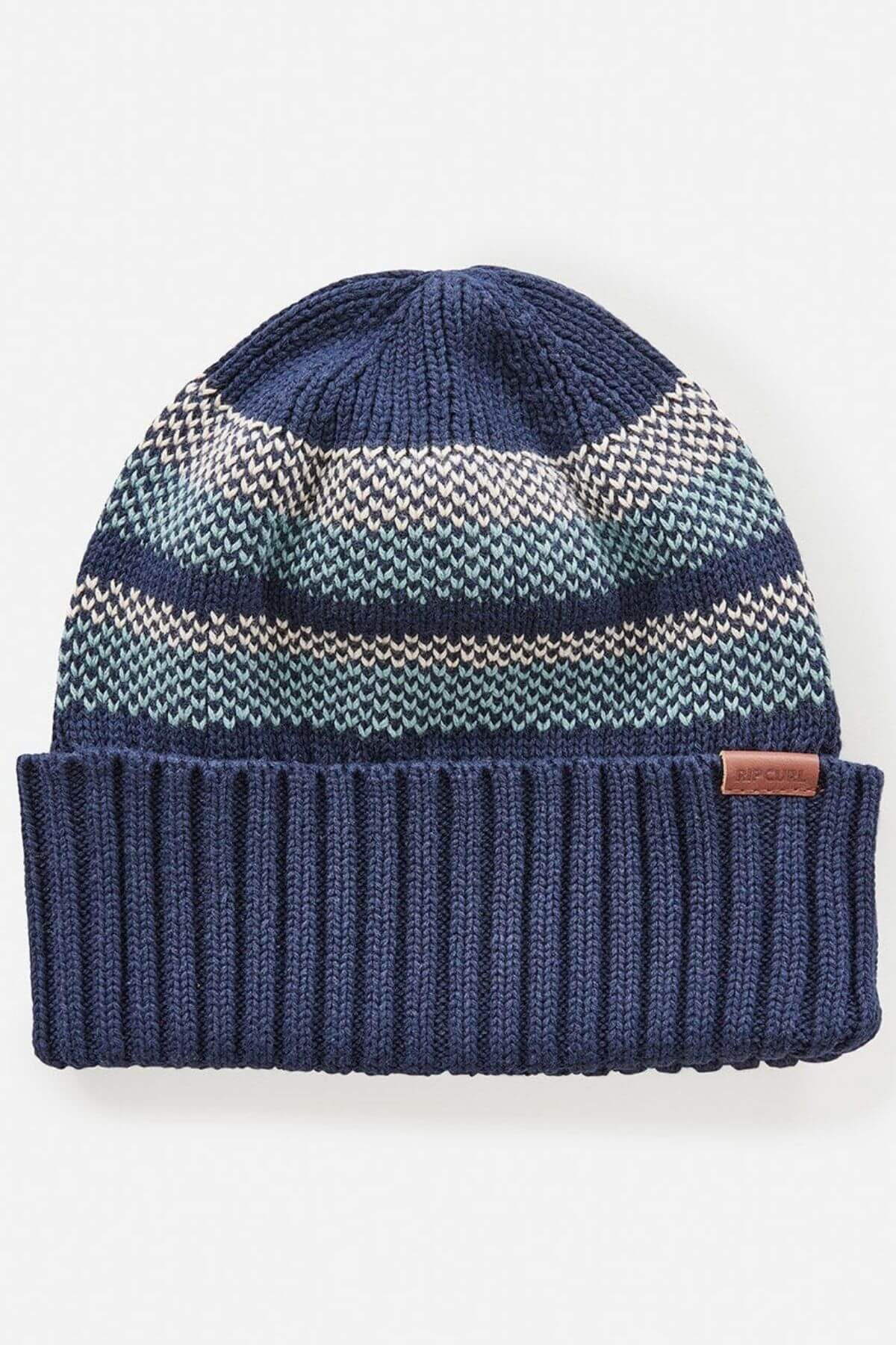 The Rip Curl Surf Revival Stripe Regular Beanie showcases a ribbed fold-over brim and a gradient pattern of white, teal, and blue stripes. A small leather patch on the brim adds style against the simple white backdrop, making it a trendy accessory.