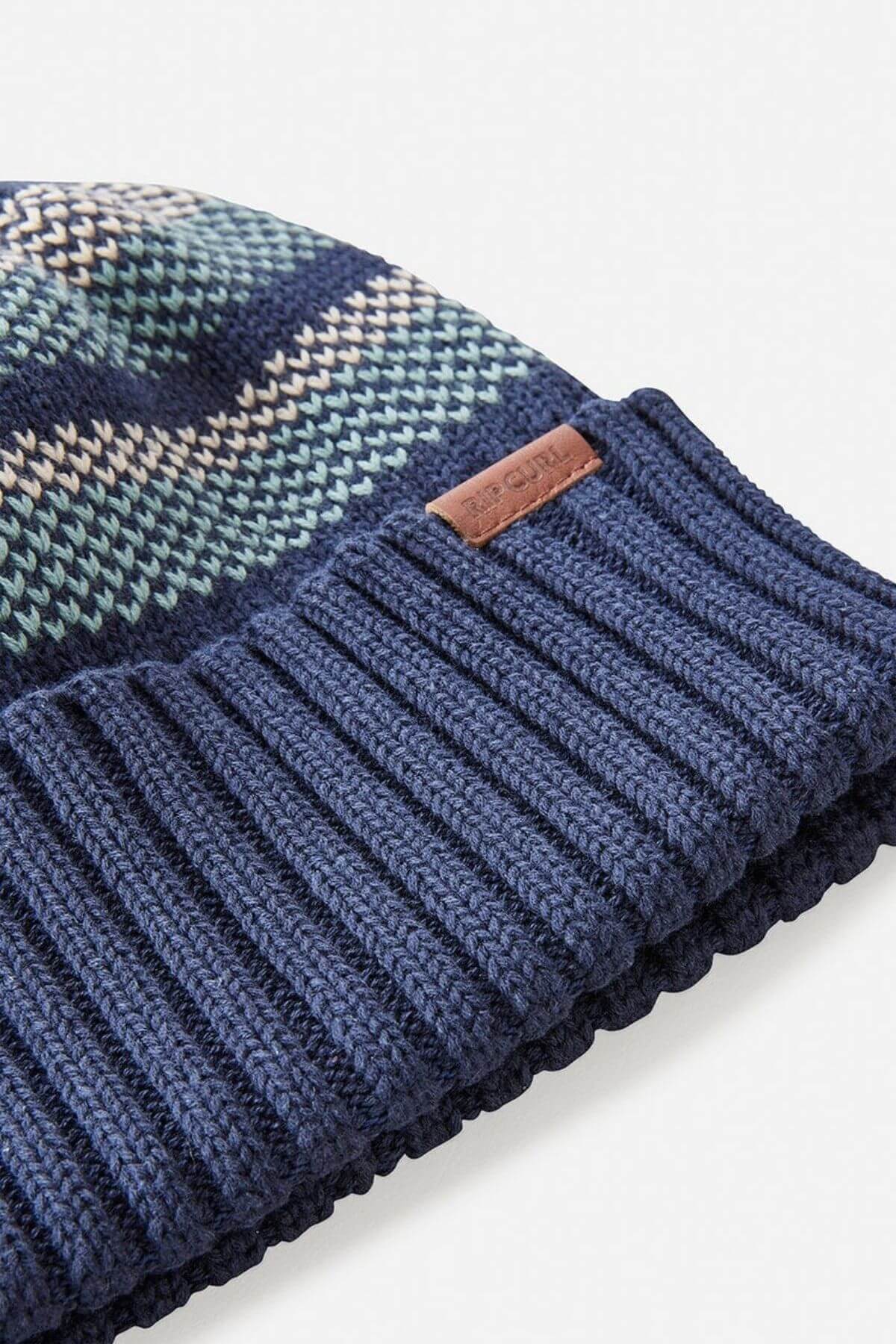 The Rip Curl Surf Revival Stripe Regular Beanie features a close-up of knitted blue, green, and beige stripes. Its bottom edge is solid navy with a ribbed texture and includes a small brown leather patch.