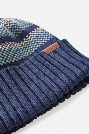 The Rip Curl Surf Revival Stripe Regular Beanie features a close-up of knitted blue, green, and beige stripes. Its bottom edge is solid navy with a ribbed texture and includes a small brown leather patch.