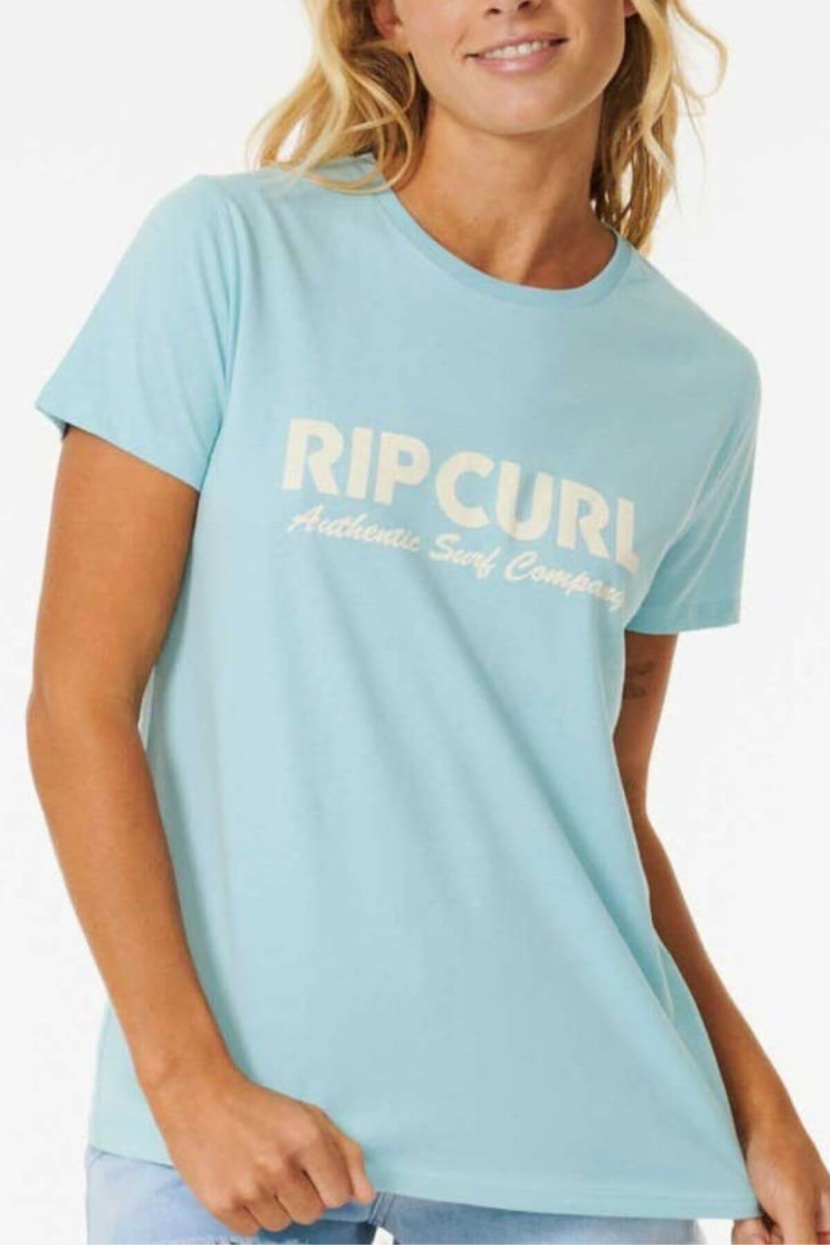 A woman wears the Rip Curl Surf Spray Standard Tee, a light blue cotton jersey with "RIP CURL Authentic Surf Company" in white on the front—ideal for relaxing by the beach and catching waves.
