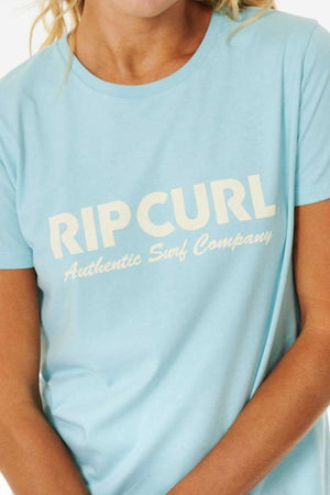 A person wearing the Rip Curl Surf Spray Standard Tee—a light blue cotton jersey t-shirt with "RIP CURL Authentic Surf Company" printed in white on the front. Only their upper torso and part of their face are visible.