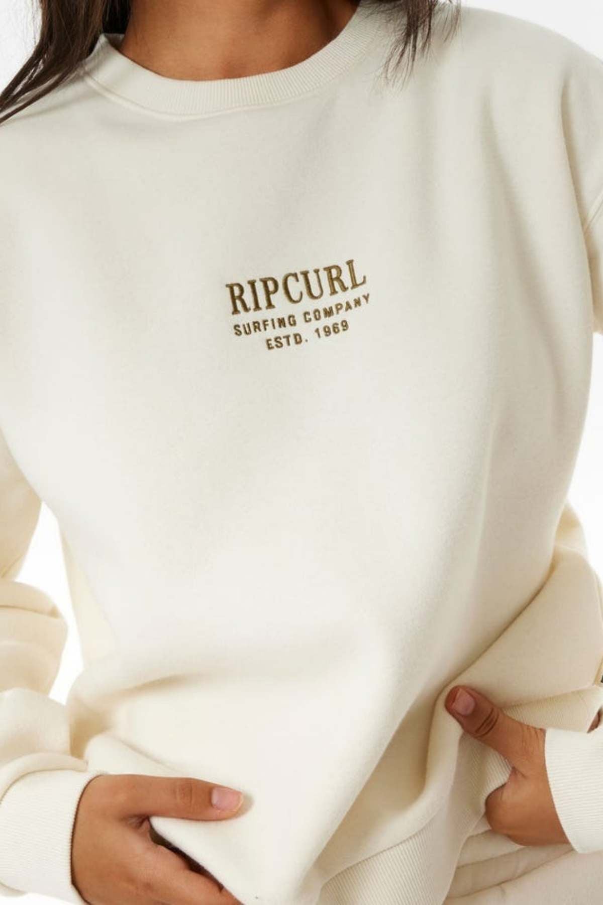 Wearing a cream Rip Curl Surf Staple Relaxed Crew featuring "RIP CURL SURFING COMPANY ESTD. 1969" in gold on the chest, a person enjoys the cozy fleece lining while holding the bottom of the sweatshirt with both hands.
