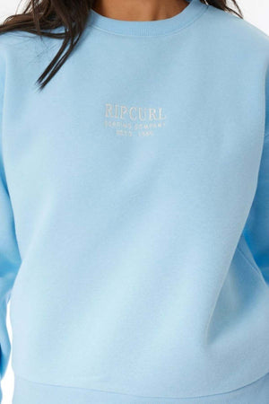 A person wears a light blue Rip Curl Surf Staple Relaxed Crew sweatshirt with the "Rip Curl Surfing Company Estd. 1969" logo in white on the chest; only their upper torso and part of their hair are visible.