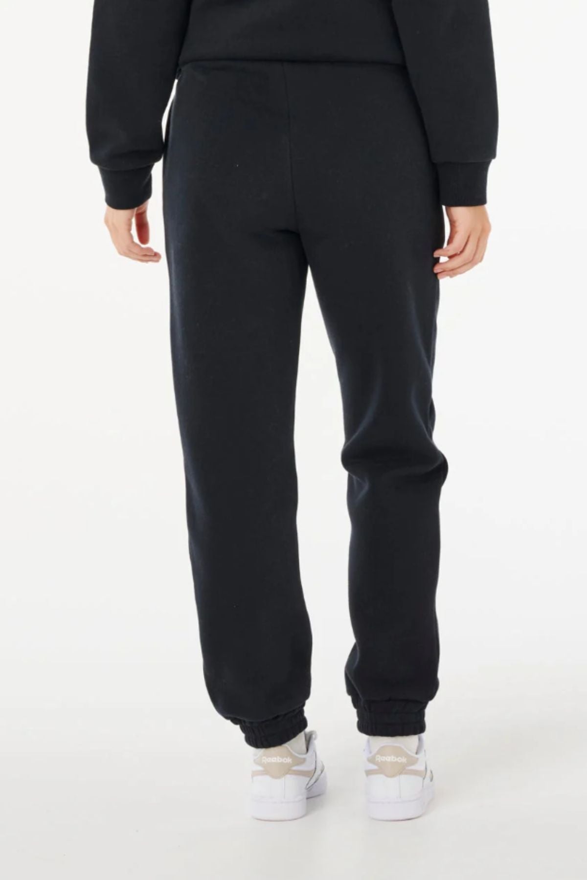 A person is standing with their back to the camera, wearing black Rip Curl Surf Staple Trackpant, a matching black sweatshirt, and white sneakers against a plain white background.