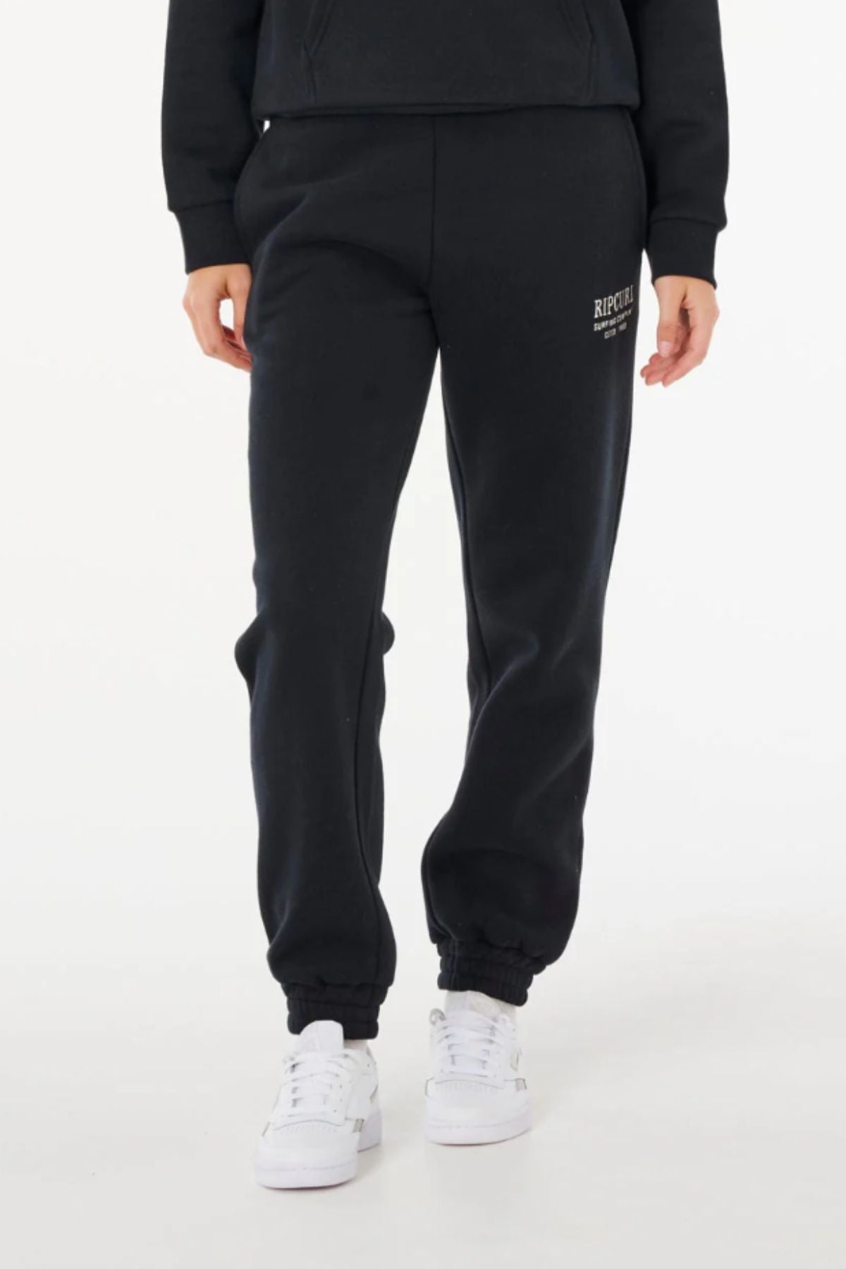 A person wears black Rip Curl Surf Staple Trackpant with an elastic waist and cuffs, a small logo on the upper left thigh, paired with white sneakers, standing against a plain white background.