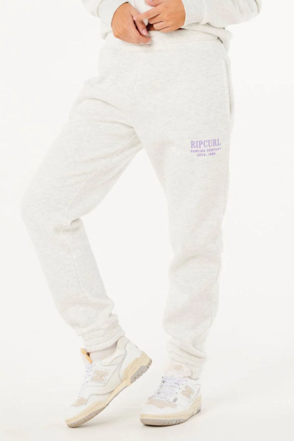 Wearing the Rip Curl Surf Staple Trackpant in light gray with purple text, paired with a matching sweatshirt and white sneakers, someone poses against a white background—ideal for relaxed, casual style.