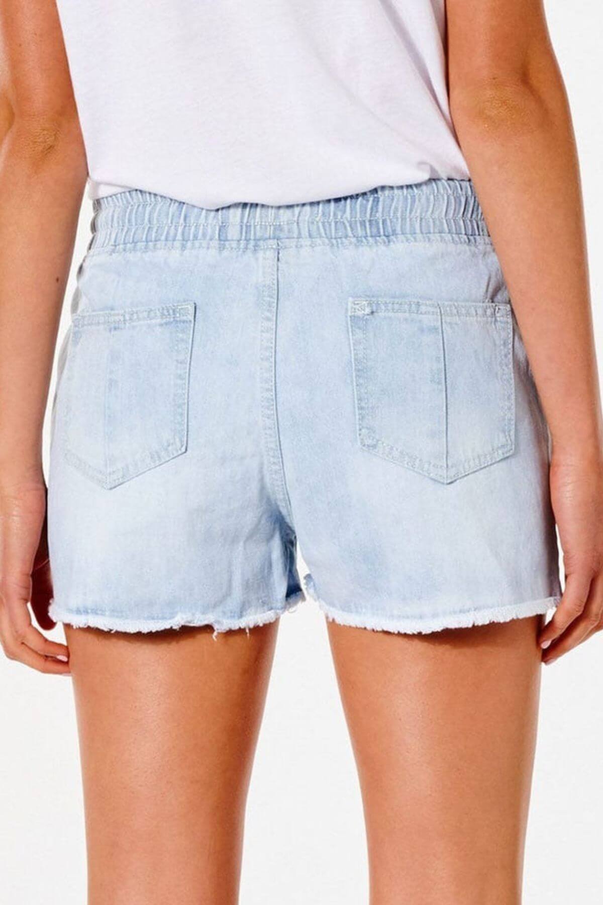 A person in a white top is seen from behind wearing Rip Curl Tara Shorts, featuring light blue denim, an elastic waistband, and two back pockets. Their arms naturally rest at their sides against a pristine white background.
