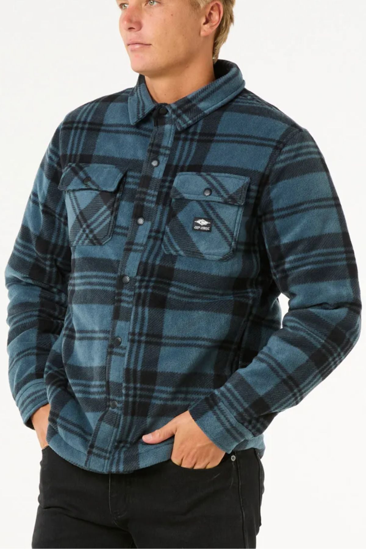 A person wearing the Rip Curl Tofino Polar Fleece Jacket in blue and black, made from recycled poly fleece with front pockets, stands with one hand in their pocket, looking off to the side against a plain white background.