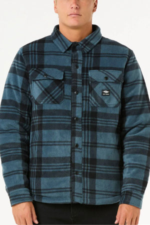 A person wears a Rip Curl Tofino Polar Fleece Jacket in blue and black plaid with chest pockets, made from recycled poly fleece, standing against a plain white background.