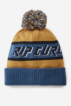 The Rip Curl Tow In Tall Beanie, in blue and mustard yellow, is made from acrylic yarn and features a playful pom-pom. It has blue cuffs and a stripe with bold "RIP CURL" lettering, making it a trendy addition to your winter wardrobe.