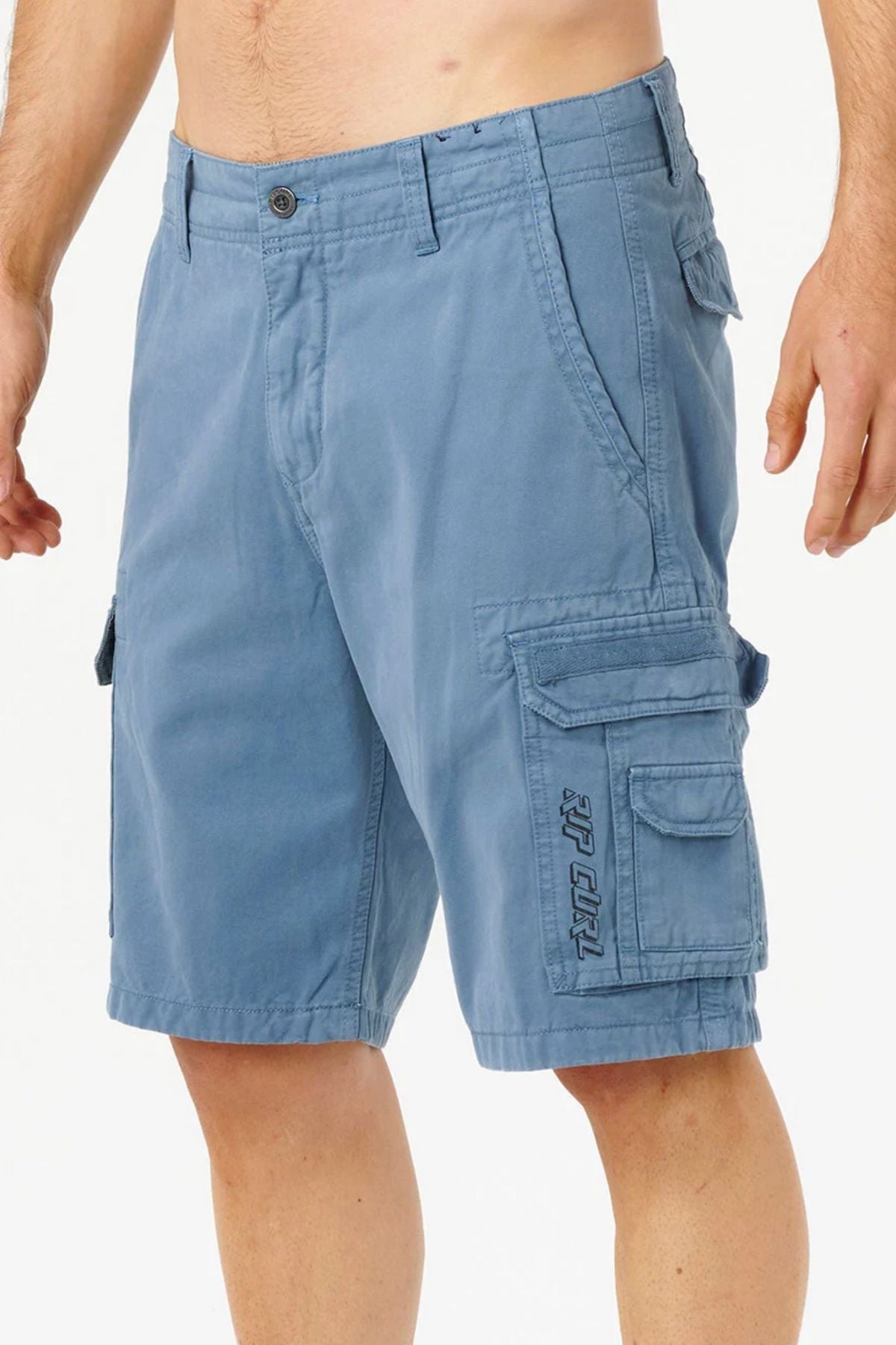 A man is wearing Rip Curl Classic Surf Trail Cargo Shorts in blue cotton with a relaxed fit, multiple pockets, button closure, decorative stitching, and an embroidered logo on the left pocket. Only his torso and legs are visible against a plain background.