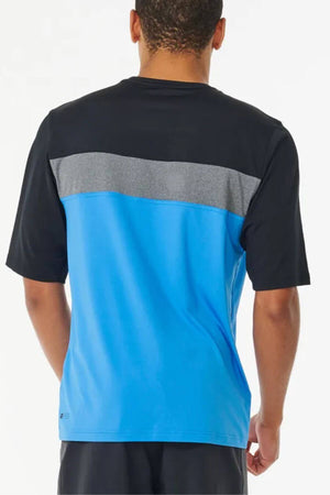 A man is seen from behind wearing the Rip Curl Undertow Short Sleeve UV Tee, which has a black top, gray stripe, and bright blue lower section. The UPF50+ tee provides extra sun protection, paired with black shorts.