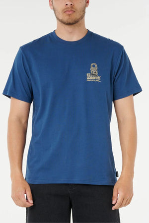 A person wearing a Rip Curl Vaporcool Search Stack Tee in blue with a small graphic and text on the left chest, paired with black pants, stands against a plain white background.