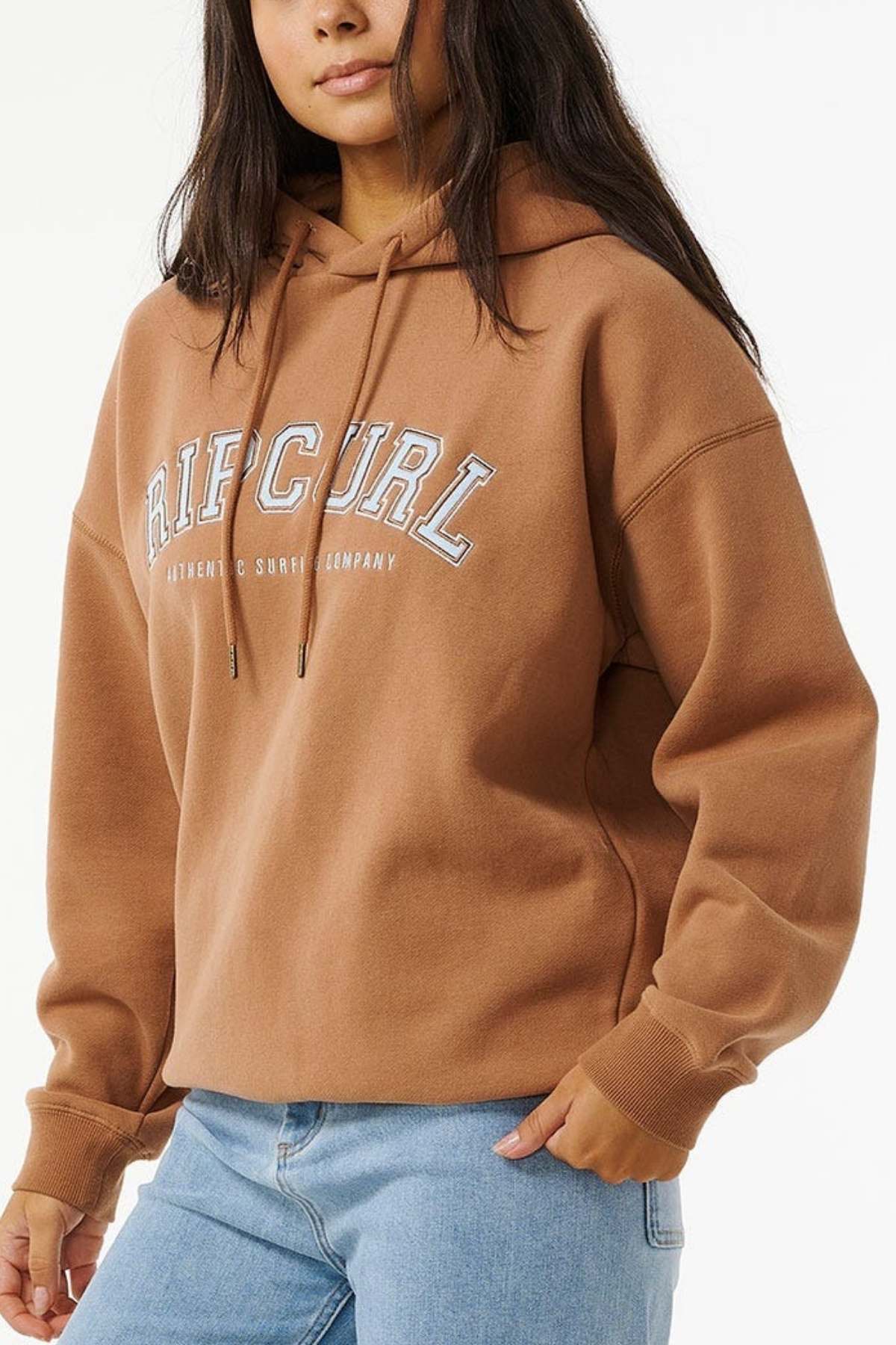 Wearing a Rip Curl Varsity Hoodie that offers an oversized fit and soft fleece lining, a person in light blue jeans strikes a confident pose with one hand on their hip against a plain white background.