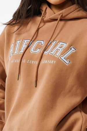 Someone is wearing a Rip Curl Varsity Hoodie in brown, featuring "Authentic Surfing Company" printed in white on the front. It has visible drawstrings and an oversized fit with a soft fleece lining, all against a plain white background.