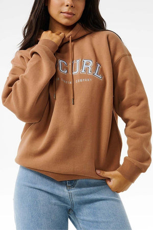A person in a brown Rip Curl Varsity Hoodie with "CURL" on it and light blue jeans stands relaxed against a plain background. The hoodie, with an adjustable drawstring hood, offers an oversized fit and soft fleece lining for extra comfort.
