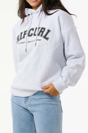 A person wears a light gray Rip Curl Varsity Hoodie with black lettering and a relaxed fleece-lined fit, paired with light blue jeans. Their long brown hair is visible against a plain white background.
