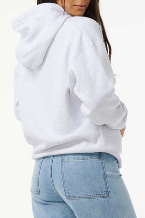 A woman models the Rip Curl Varsity Hoodie in an oversized light gray fit with light blue jeans, standing with her back partially turned and looking over her shoulder against a plain white background.