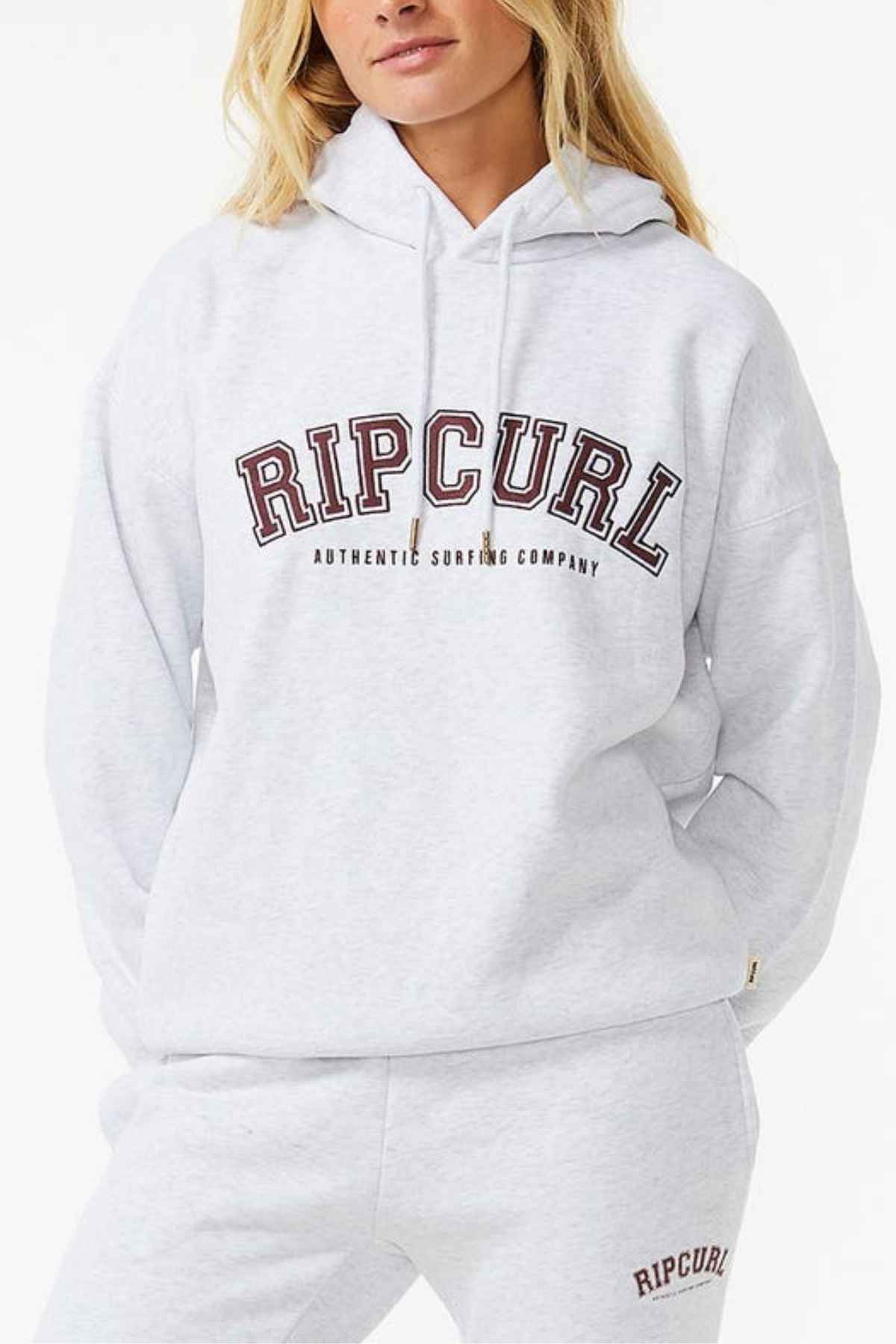 Wearing a gray Rip Curl Varsity Hoodie and sweatpants labeled "RIPCURL AUTHENTIC SURFING COMPANY," this casual set is made from Cotton Poly Fleece. The Heritage fit combines comfort and style, highlighted against a plain white background.