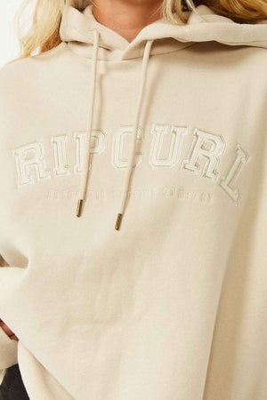 A person wears the Rip Curl Varsity Hoodie, heritage fit, made from Cotton Poly Fleece, with "RIP CURL" and "AUTHENTIC SURFING COMPANY" embroidered in white on the front. Only their lower face, torso, and part of hair are visible.