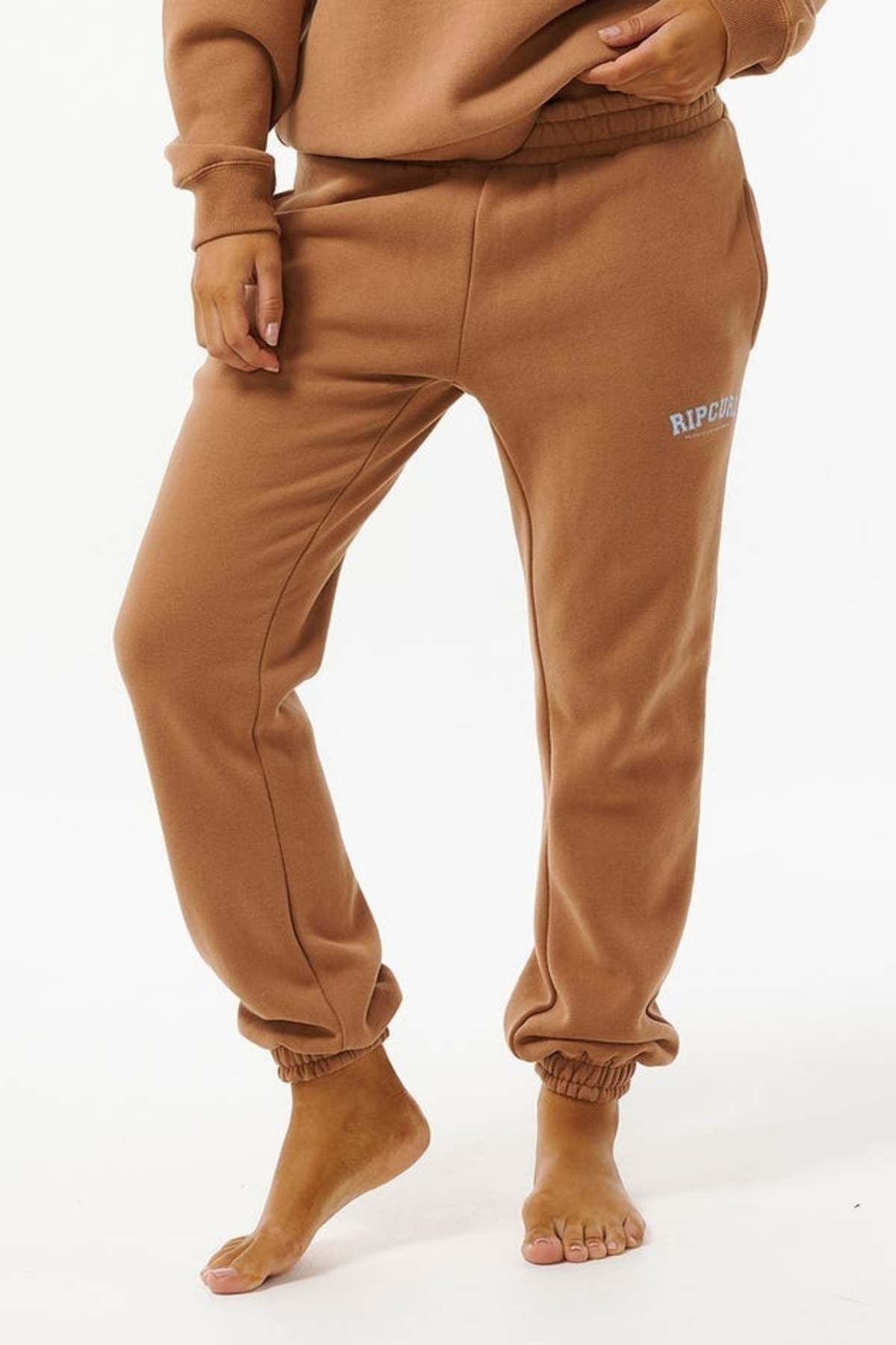 A person in Rip Curl Varsity Pants, crafted from a cotton/polyester blend with elastic waistband and cuffs, stands barefoot on a white background. "RIP CURL" branding appears on the left thigh, combining comfort and style.