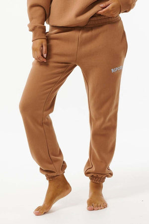 A person in Rip Curl Varsity Pants, crafted from a cotton/polyester blend with elastic waistband and cuffs, stands barefoot on a white background. "RIP CURL" branding appears on the left thigh, combining comfort and style.