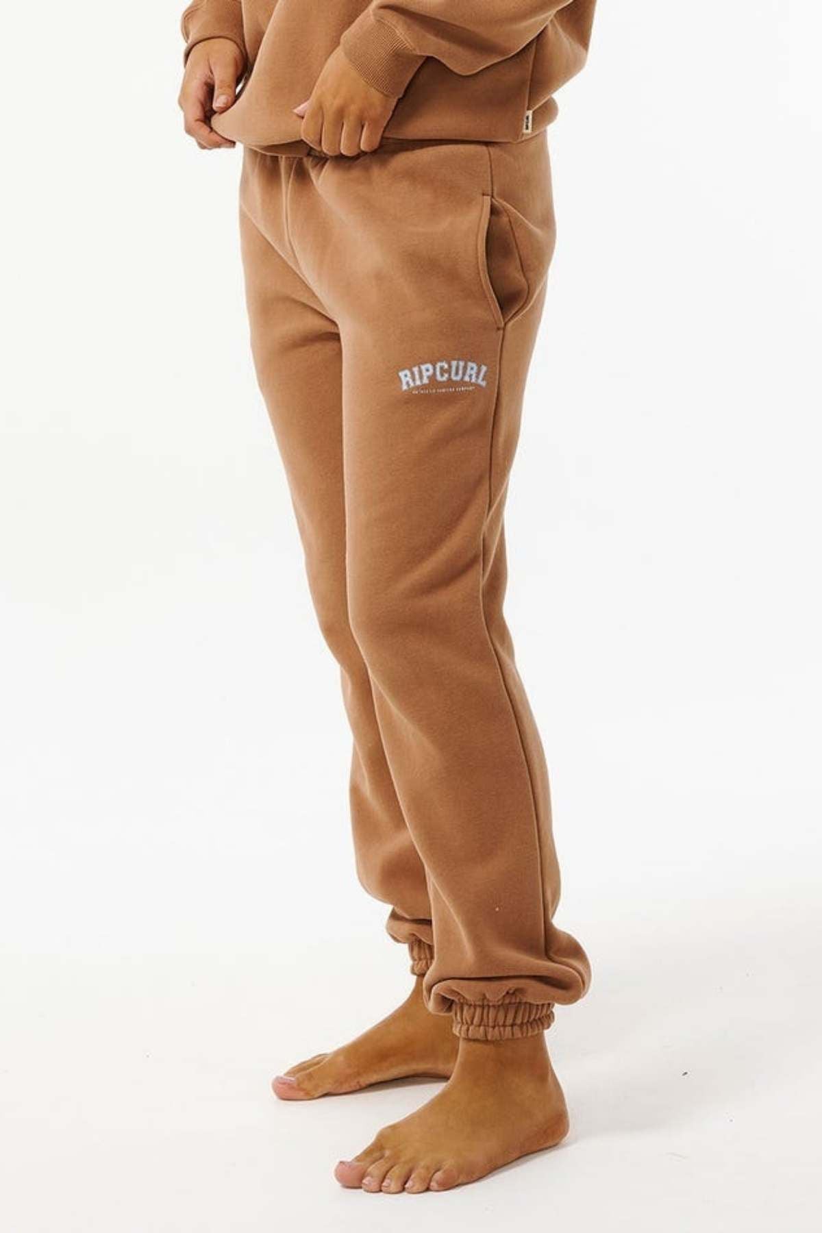 Wearing Rip Curl Varsity Pants made from a soft cotton/polyester blend with "RIP CURL" on the left thigh, a person stands barefoot against a white background, showcasing the pants' comfort and style.