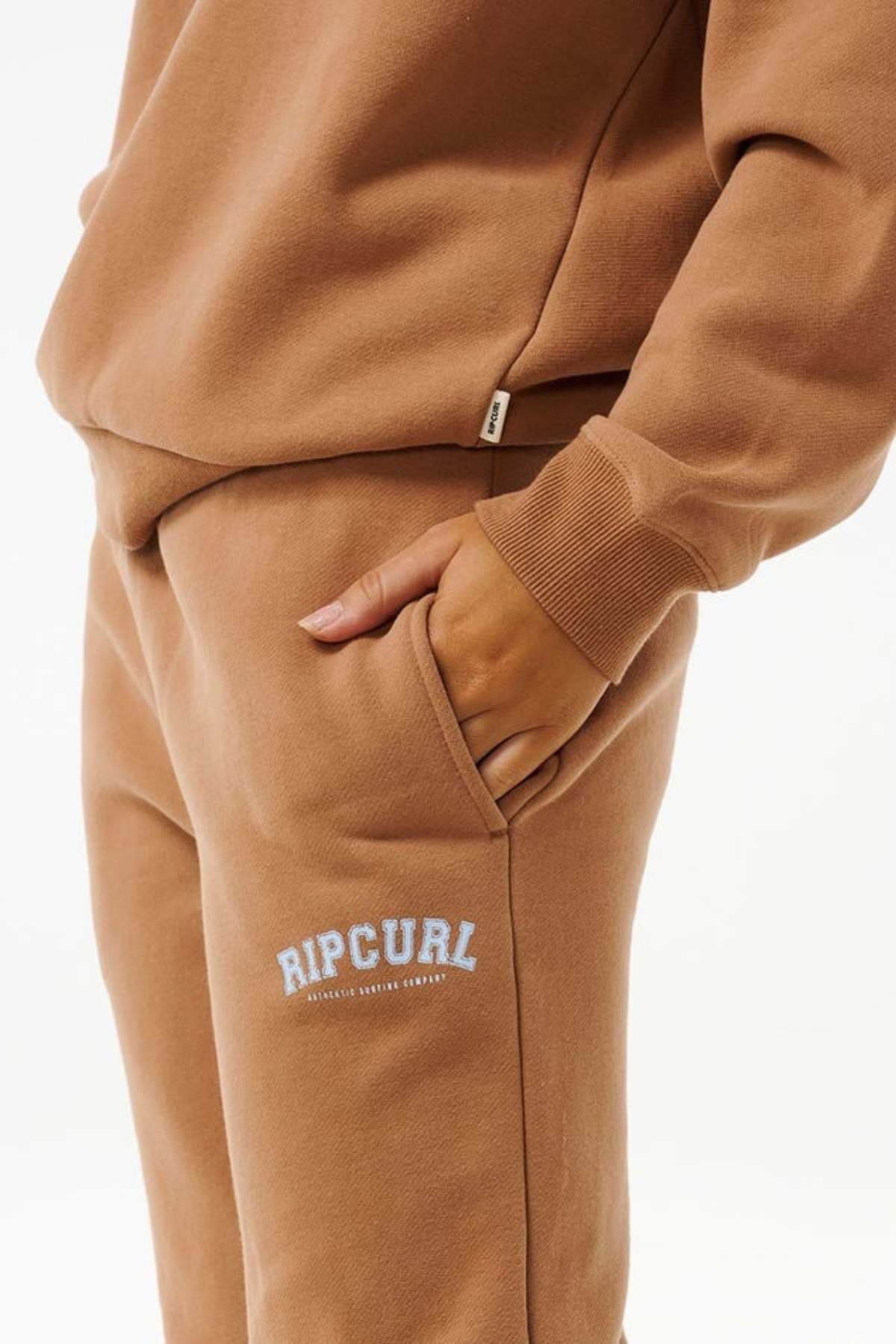 A person wears Rip Curl Varsity Pants, made from a Cotton/Polyester Blend, with one hand in their pocket; the sweatpants feature a white "RIP CURL" logo near the pocket for comfort and style.