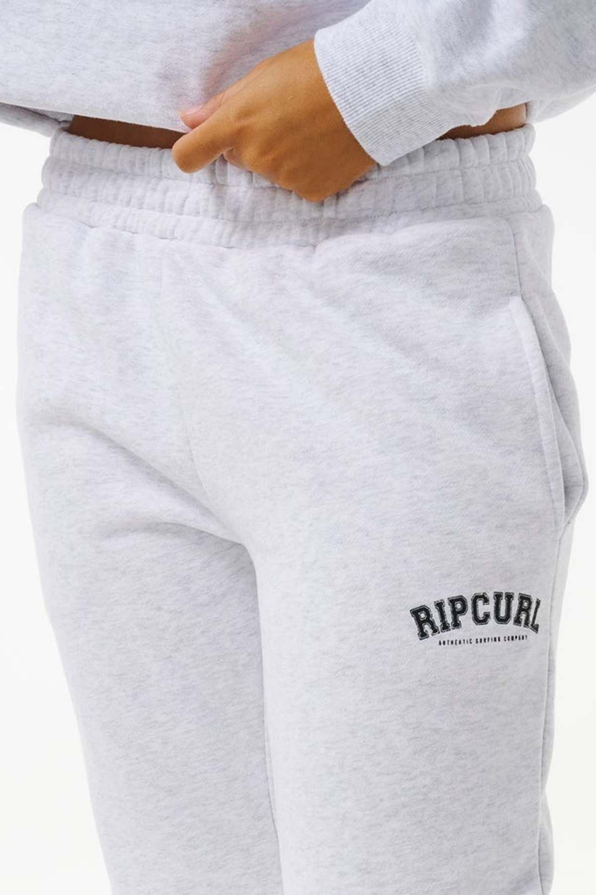 A person in casual wear dons light grey Rip Curl Varsity Pants made from Cotton Poly Fleece, with "RIP CURL" embroidered in black on the left thigh, paired with a matching sweatshirt and one hand in their pocket.