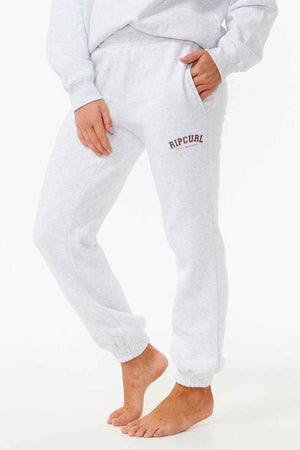 A person stands barefoot in Rip Curl Varsity Pants, crafted from soft Cotton Poly Fleece. The casual wear has an elastic waistband and cuffs with "Rip Curl" on the thigh. One hand is in their pocket, set against a plain white background.