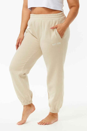 A person wears beige Rip Curl Varsity Pants made from Cotton Poly Fleece, with an elastic waistband and cuffs. The pants have side pockets and "RIP CURL" on the upper thigh, shown barefoot against a white backgroundโideal for casual wear.