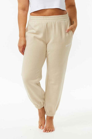 Wearing Rip Curl Varsity Pants in light beige fleece and a white cropped top, a barefoot model stands against a plain background with hands by their sides and one leg bent, highlighting the effortless casual style of the pants.