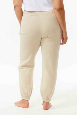 A person stands barefoot facing away, wearing Rip Curl Varsity Pantsโa beige jogger-style sweatpant made from soft cotton poly fleeceโwith a white top, set against a plain white background to highlight this casual outfit.