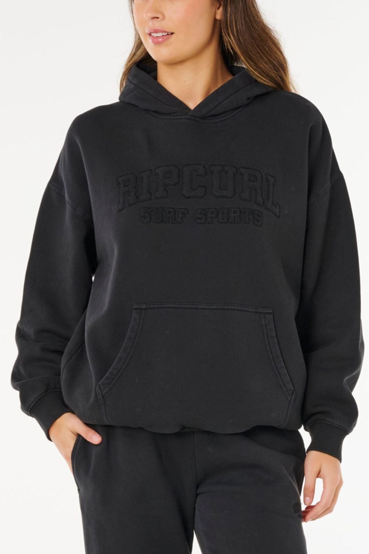 A person is wearing a black Rip Curl Varsity Sports Oversized Hood with a front pocket and matching black pants, standing casually with one hand in their pocket against a plain background.
