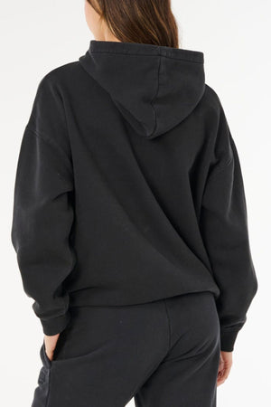 A person with long brown hair, facing away from the camera against a plain white background, wears the Rip Curl Varsity Sports Oversized Hood and black pants, showcasing an effortless casual style.