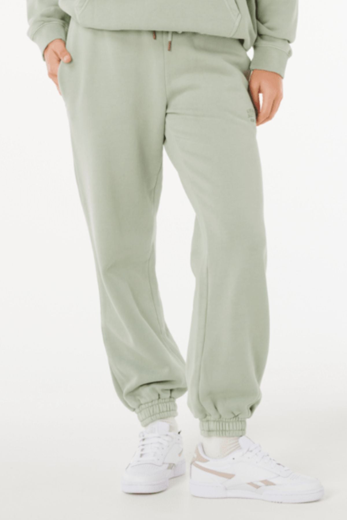 Wearing light green Rip Curl Varsity Sports Track Pant, a matching sweatshirt, and white sneakers, a person stands with hands in pockets against a plain white background.