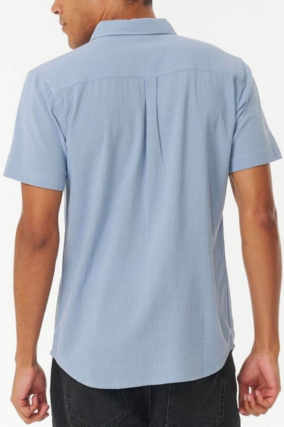 A person is shown from the back against a plain white background, wearing a light blue Rip Curl Washed Short Sleeve Shirt and dark pants.