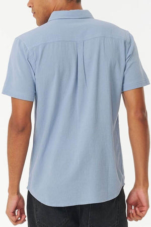 A person is shown from the back against a plain white background, wearing a light blue Rip Curl Washed Short Sleeve Shirt and dark pants.