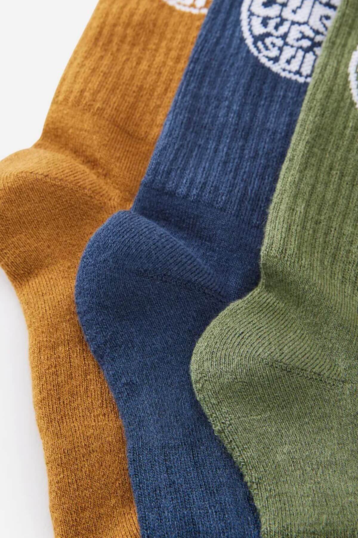 Close-up of three overlapping Rip Curl Wetty Crew socks in ochre, navy blue, and olive green on a white background. Each ribbed sock has a white circular logo near the top and provides arch support.