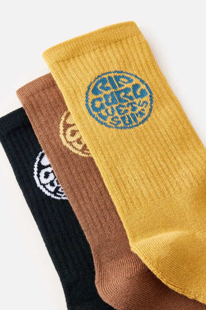 Three pairs of socks from the Rip Curl Wetty Crew Sock 3 Pack are displayed, each laid one over the other. The top pair is yellow with a blue circular emblem, the middle pair is brown with a yellow emblem, and the bottom pair is black with a white emblem. Each sock promises arch support and ultimate comfort, featuring stylized text designs on their emblems.