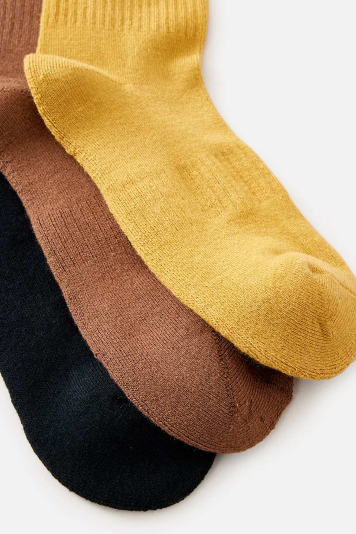 Three pairs of socks from the Rip Curl Wetty Crew Sock 3 Pack are arranged in a fan shape, showcasing ultimate comfort with their ribbed texture at the cuff. The mustard yellow Wetty Crew Sock is positioned on top of the brown and black pairs beneath it, all crafted from the same fabric that features arch support for enhanced wearability.